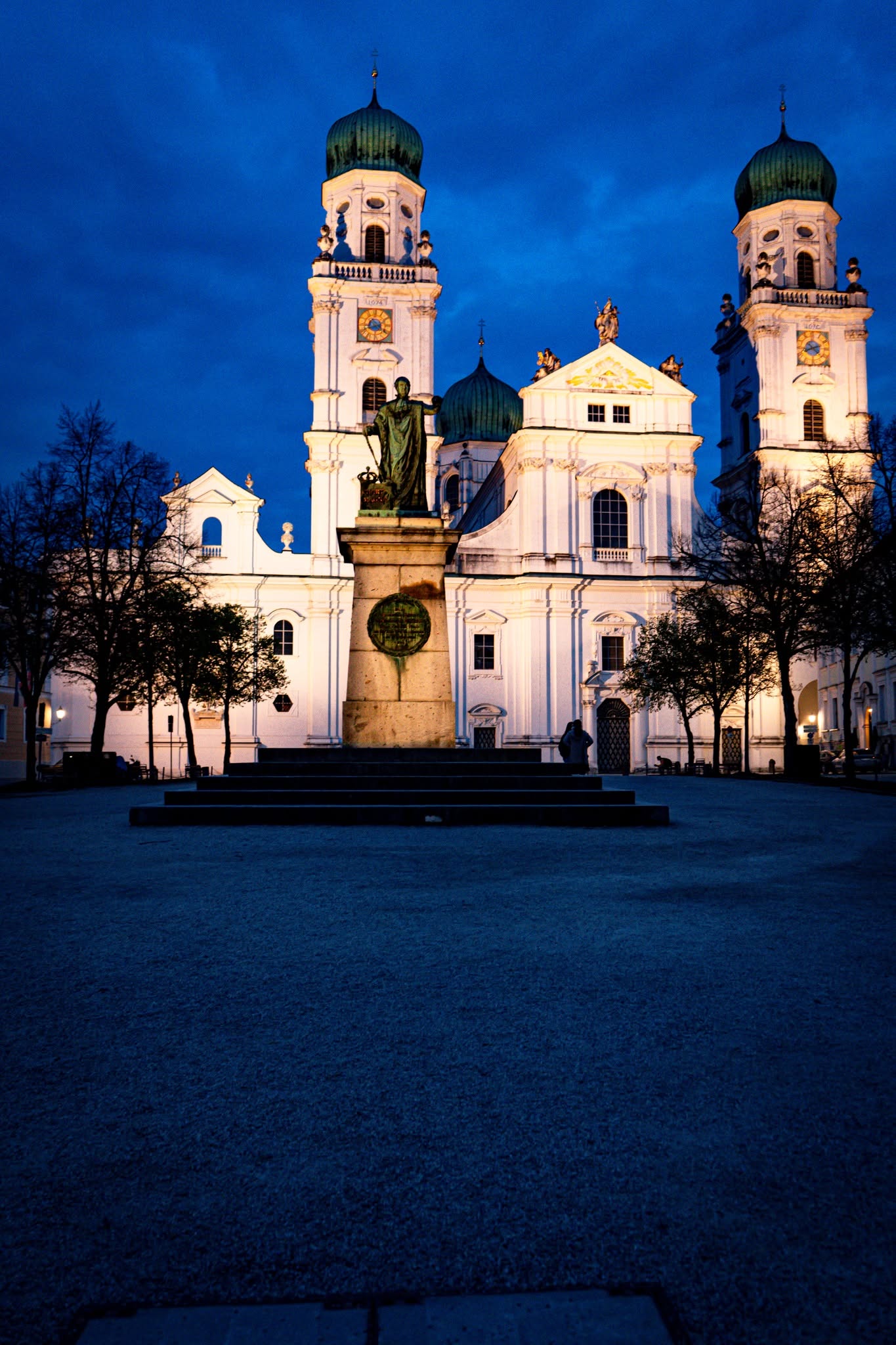 Passau Dom St. Stephan bei Nacht — beleuchtete barocke Fassade mit Statue und grünen Kuppeln vor blauem Abendhimmel