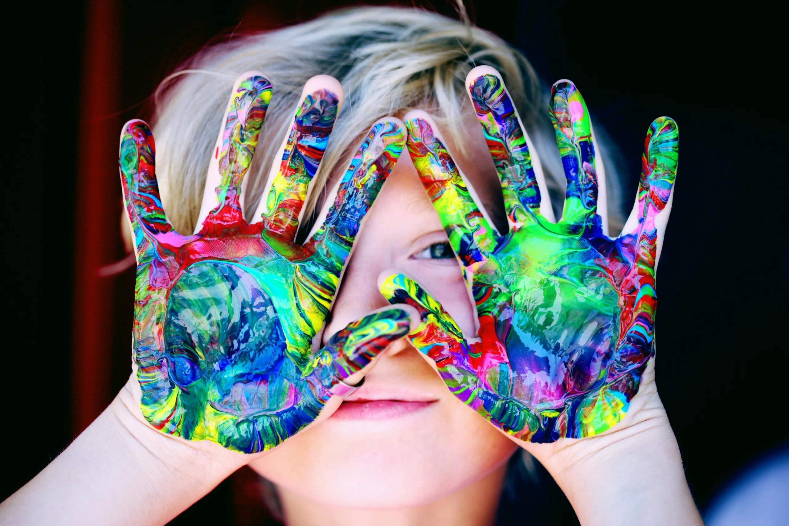 Child with colourful painted hands peeking through fingers