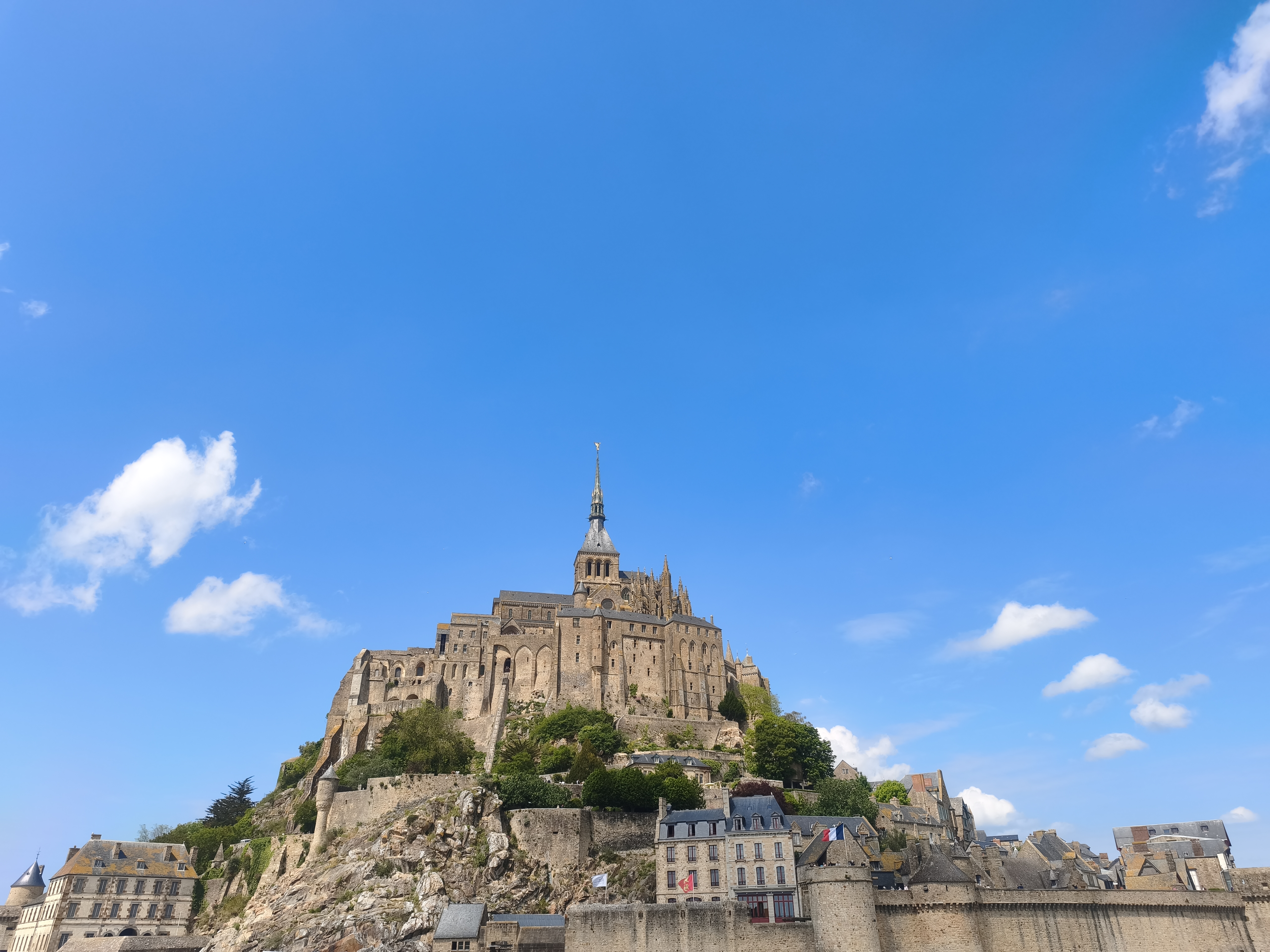 Mont-Saint-Michel rising above the flat Normandy coast under a bright blue sky with scattered white clouds